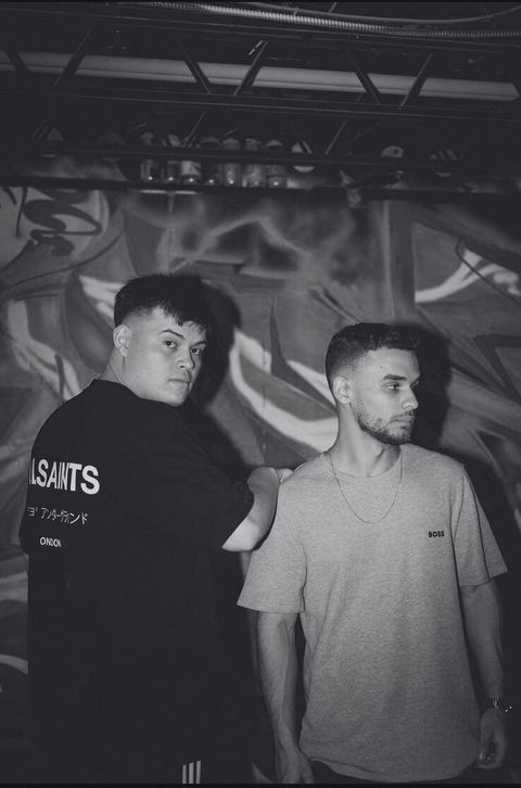 Black and white portrait of the duo in front of a graffiti wall.